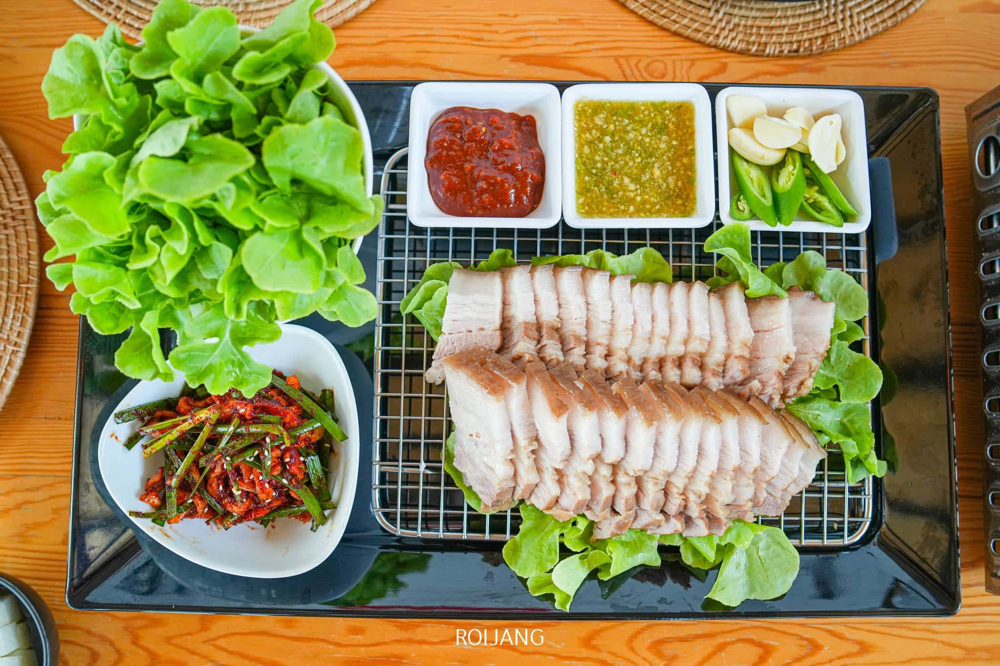 หมูสามชั้นต้มหั่นเป็นแผ่นบนเตียงผักใบเขียว พร้อมด้วยผักกาดหอม กระเทียม พริกเขียวหั่นเป็นชิ้น น้ำจิ้ม 3 อย่าง และเครื่องเคียงผักรวมรอคุณอยู่ที่ร้านอาหารเกาหลีภูเก็ต