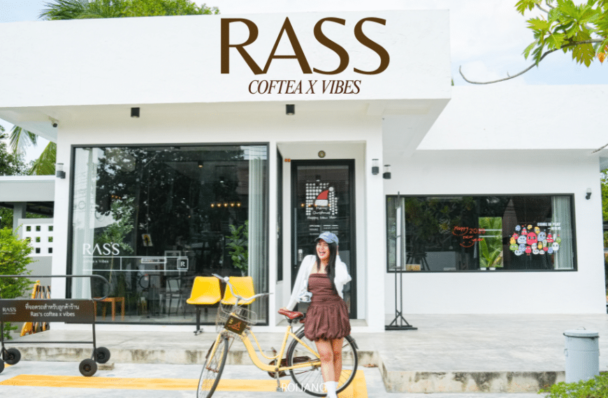 หญิงสาวกับจักรยานยืนอยู่หน้า "Ras's Coftea X Vibes" คาเฟ่โคกกลอยอันมีเสน่ห์ที่มีประตูกระจก เก้าอี้สีเหลืองสดใส และต้นไม้เขียวขจี.