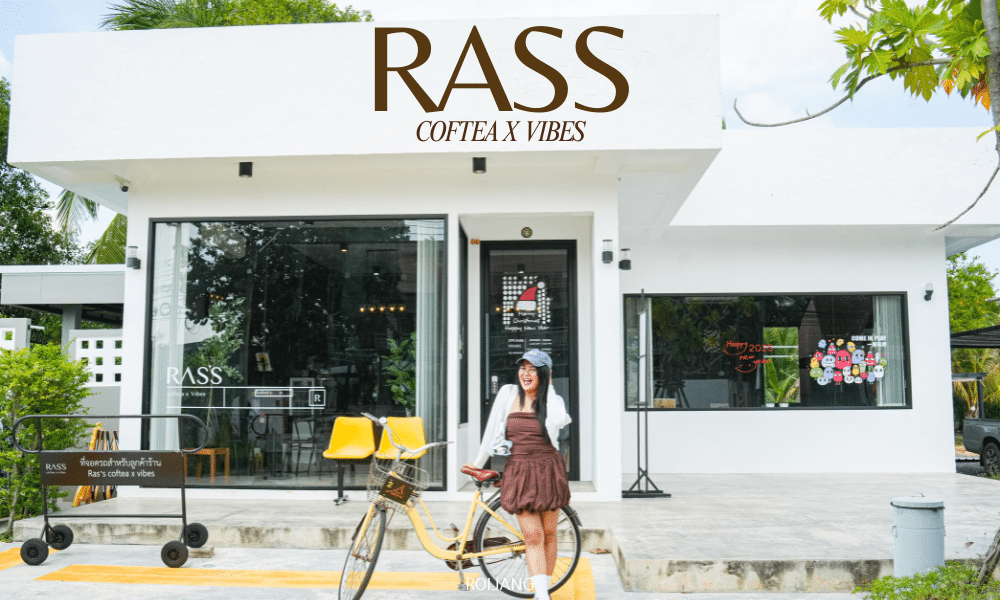 หญิงสาวกับจักรยานยืนอยู่หน้า "Ras's Coftea X Vibes" คาเฟ่โคกกลอยอันมีเสน่ห์ที่มีประตูกระจก เก้าอี้สีเหลืองสดใส และต้นไม้เขียวขจี.