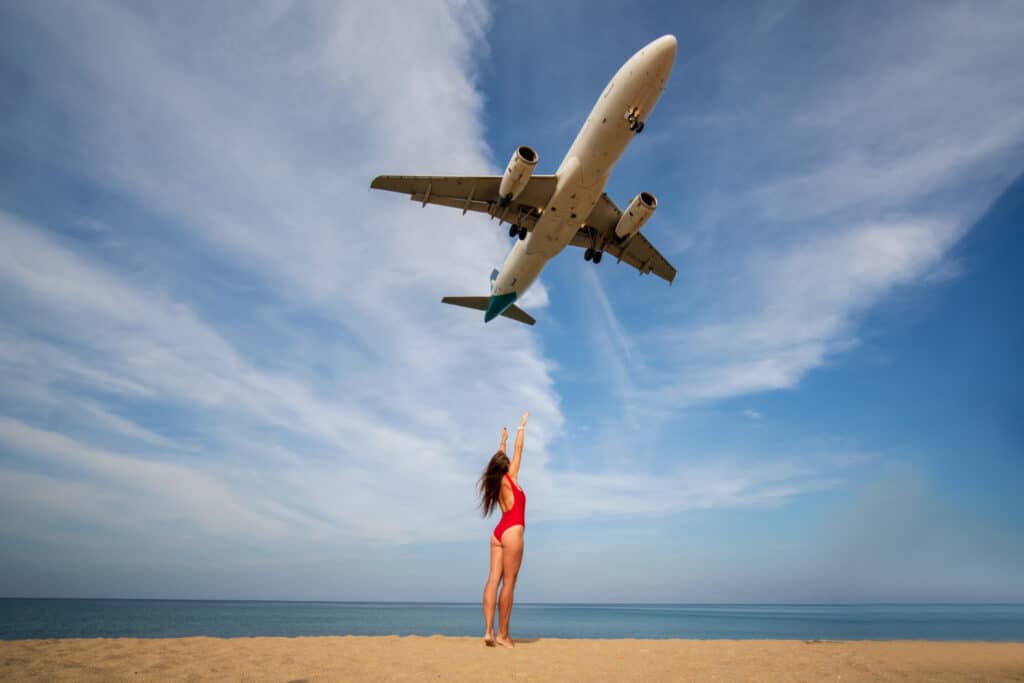 ผู้หญิงในชุดว่าย ที่เที่ยวภูเก็ต น้ำสีแดงยืนอยู่บนชายหาดโดยมีเครื่องบินบินอยู่เหนือศีรษะ