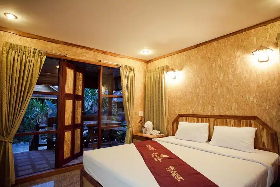ห้องพักในโรงแรมที่มีเตียงที่ทำขึ้นอย่างประณีตและประตูกระจกบานเลื่อนในที่พักของเกเมรัทธา ที่พักเขมราฐ