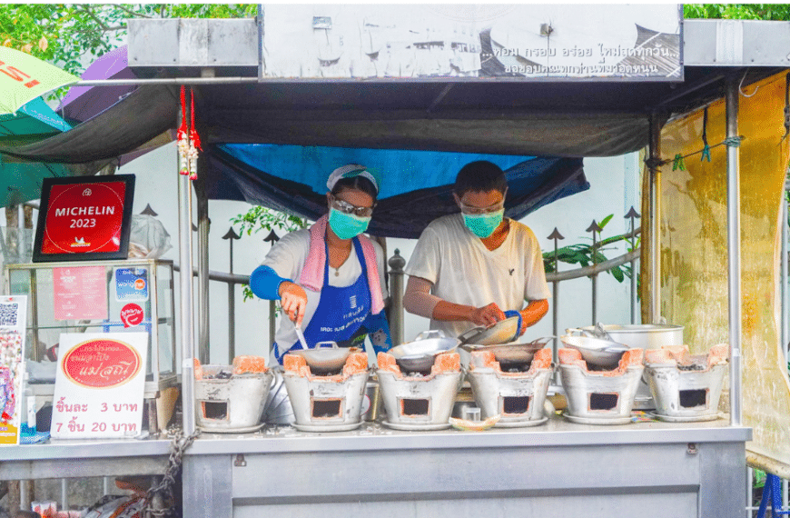 คนสองคนกำลังทำอาหารที่แผงขายอาหารในย่านเมืองเก่าอันเก่าแก่ของภูเก็ต โดยมีขนมท้องถิ่น เช่น ขนมอาโปงแม่ส