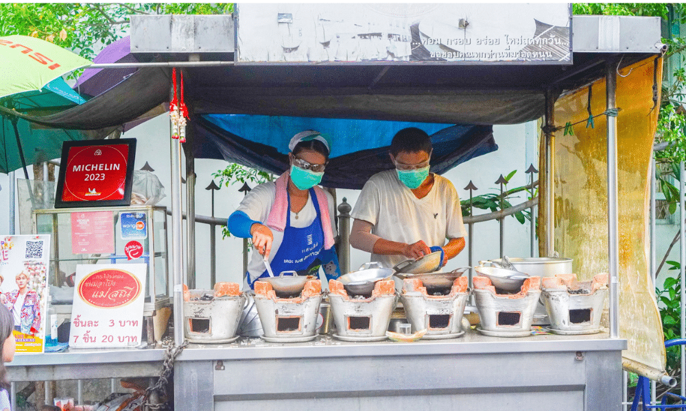 คนสองคนกำลังทำอาหารที่แผงขายอาหารในย่านเมืองเก่าอันเก่าแก่ของภูเก็ต โดยมีขนมท้องถิ่น เช่น ขนมอาโปงแม่ส