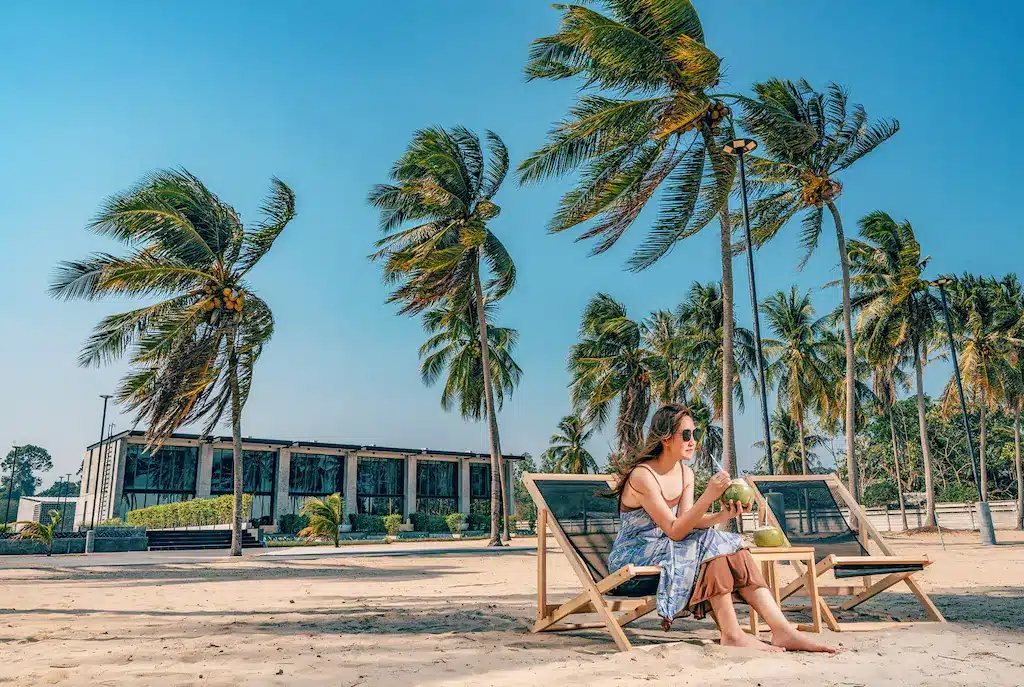 ผู้หญิงนั่งเล่นบนเก้าอี้ชายหาดในรีสอร์ทเขตร้อน โดยมีต้นปาล์มเป็นฉากหลัง ที่พักทับสะแก