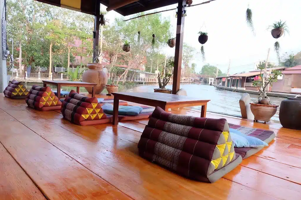 ชานไม้ที่มีโต๊ะและเก้าอี้ไม้อยู่ติดกับทางน้ำ ที่เที่ยวอัมพวา