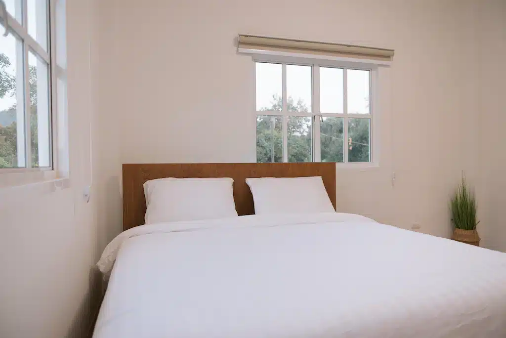 เตียงในห้องที่โรงแรมแห่งหนึ่งในสัตหีบ มีผ้าปูที่นอนสีขาวและหน้าต่าง บ้านพักสัตหีบ