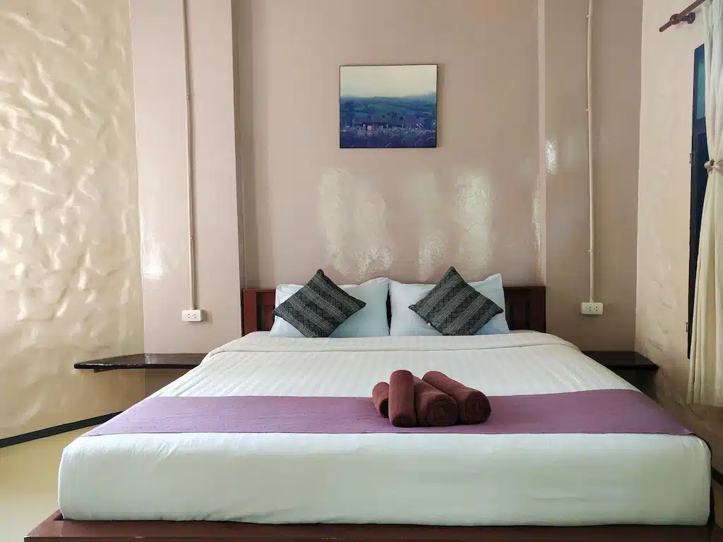 เตียงในห้องที่มีผนังสีขาวและผ้าห่มสีม่วงที่วังน้ำเขียว ที่พักวังน้ำเขียว