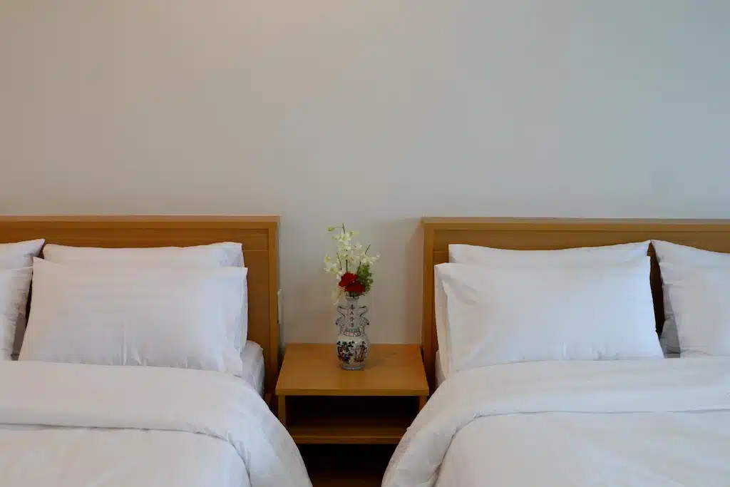 เตียงสองผืนพร้อมผ้าปูที่นอนสีขาวและแจกันดอกไม้ ณ วัดดอยแม่สลอง จังหวัดน่าน ที่พักแม่สลอง