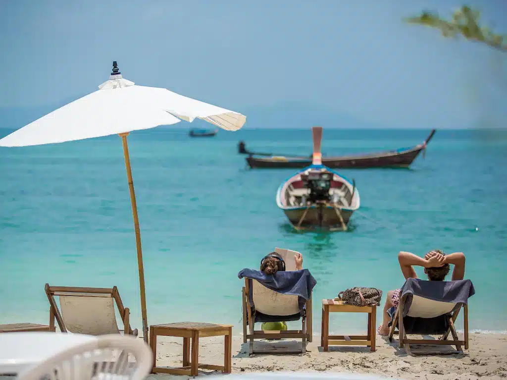 คน 2 คนกำลังนั่งอยู่บนเก้าอี้บนชายหาดเกาะกระดาน โดยมีเรือเป็นฉากหลัง ที่พักเกาะกระดาน