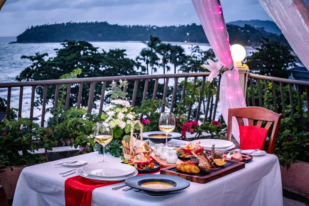 ที่ชนาลัย การ์เดน รีสอร์ท มีการจัดโต๊ะอย่างหรูหราพร้อมด้วยอาหารและไวน์ชั้นเลิศที่คัดสรรมาอย่างดีบนระเบียงที่น่าหลงใหล Chanalai garden resort พร้อมทิวทัศน์อันตระการตาของมหาสมุทร