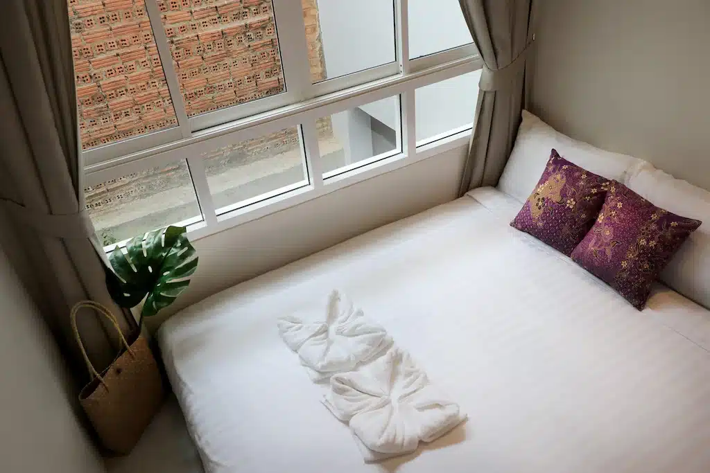 เตียงพร้อมผ้าปูที่นอนสีขาวและผ้าเช็ดตัวริมหน้าต่าง ณ ที่พักริมชายหาดในจังหวัดระนอง ที่พักระนองในเมือง