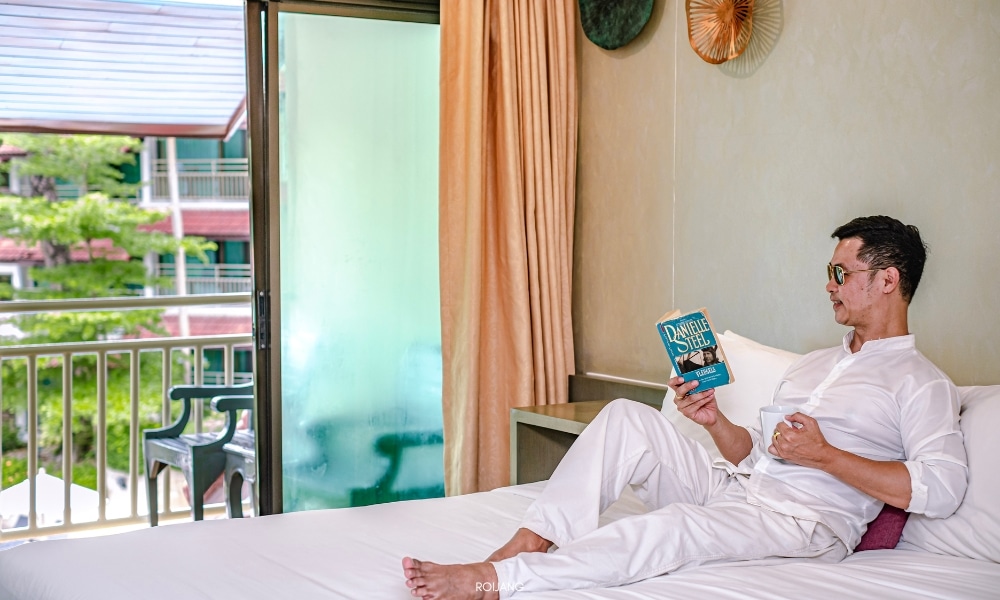 ผู้ชายกำลังอ่านหนังสือที่ชนาลัย ฟลอรา รีสอร์ท บนเตียง Chanalai Flora Resort