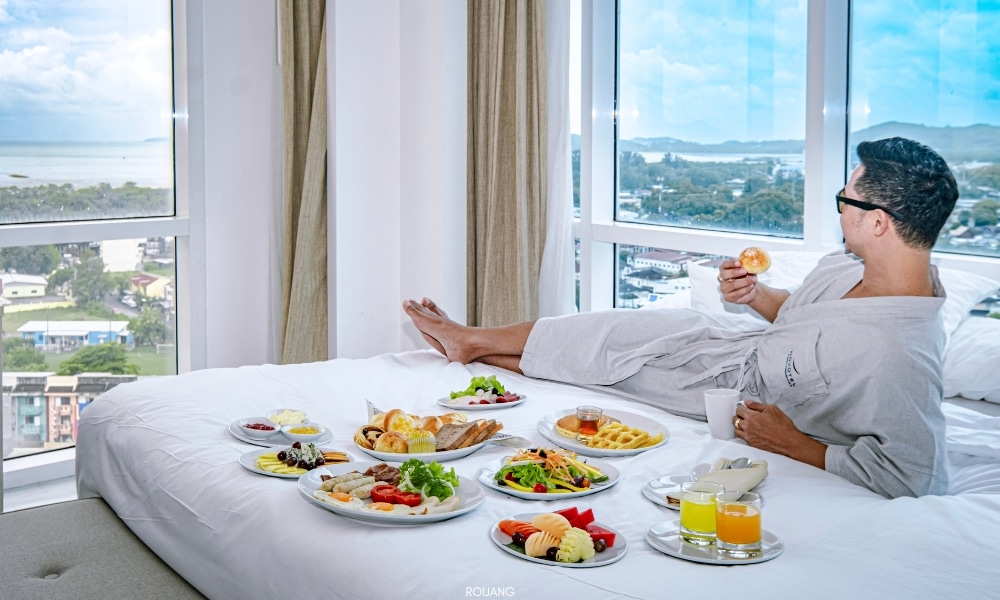 ผู้ชายกำลังนั่งอยู่บนเตียงที่โรงแรมโนโวเทล ภูเก็ต ซิตี้ โภคีธรา พร้อมจานอาหาร Novotel phokeethra Phuket