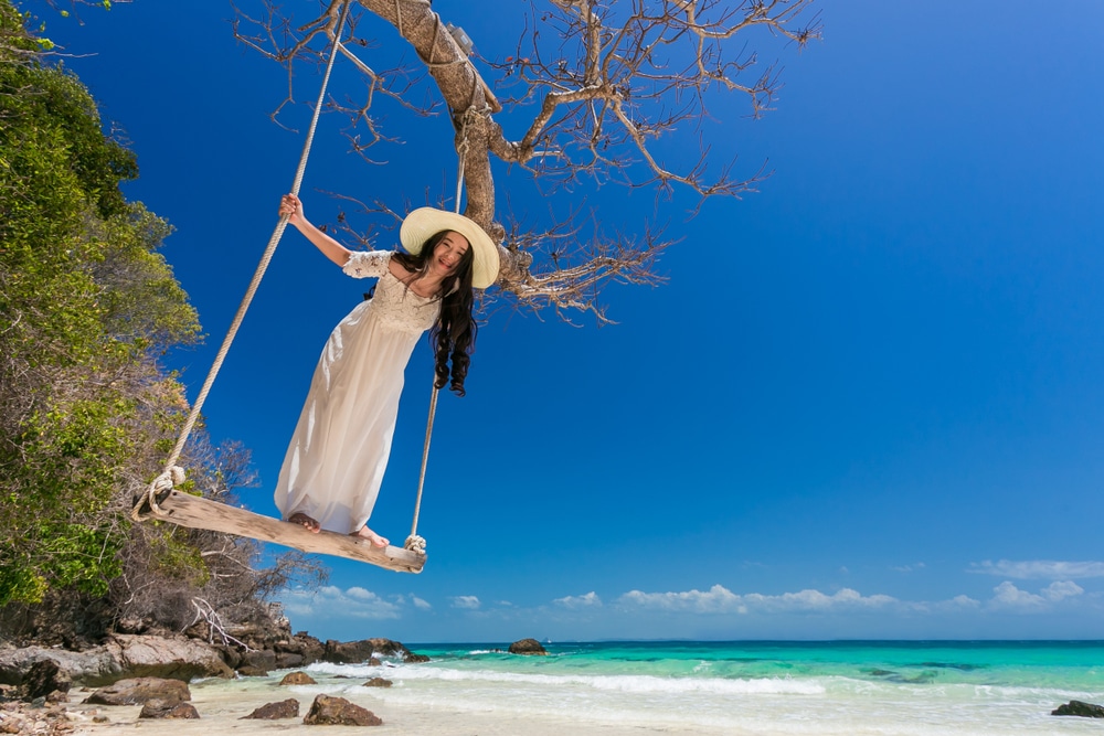       ที่เที่ยวหน้าร้อน  Description: ผู้หญิงในชุดขาวแกว่งไปมาบนต้นไม้บนชายหาดเขตร้อน เพลิดเพลินกับที่เที่ยวสงกรานต์