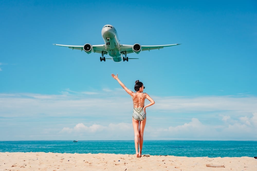 ผู้หญิงในชุดบิกินี่ยืนอยู่บนชายหาดโดยมีเครื่องบินบินอยู่เหนือศีรษะ กำลังเพลิดเพลินกับที่เที่ยวสงกรานต์ ที่เที่ยวหน้าร้อน
