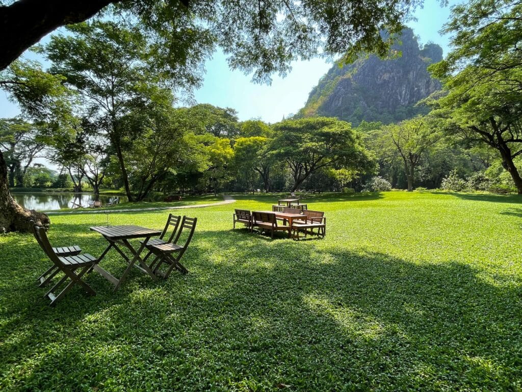สนามหญ้าที่มีโต๊ะและเก้าอี้อยู่หน้าภูเขาที่ประดับประดาด้วยต้นจามจุรียักษ์ในกาญจน ต้นจามจุรียักษ์กาญจนบุรี