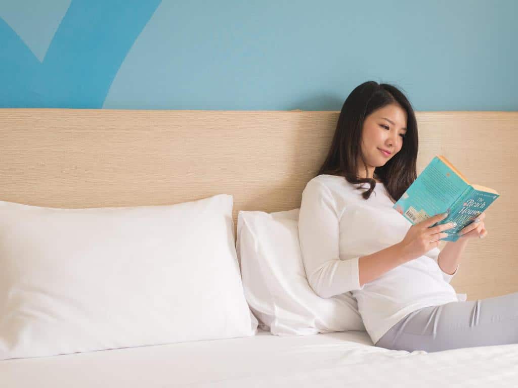 ผู้หญิงคนหนึ่งนั่งอยู่บนเตียงในโรงแรมนครราชสีมา กำลังอ่านหนังสือ ที่พักนครราชสีมา