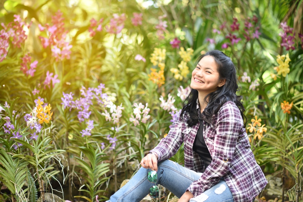 หญิงสาวนั่งอยู่บนพื้นในสวนที่เต็มไปด้วยดอกไม้ เพลิดเพลินกับทิวทัศน์ที่สวยงามของราชบุรี เที่ยวสวนผึ้ง