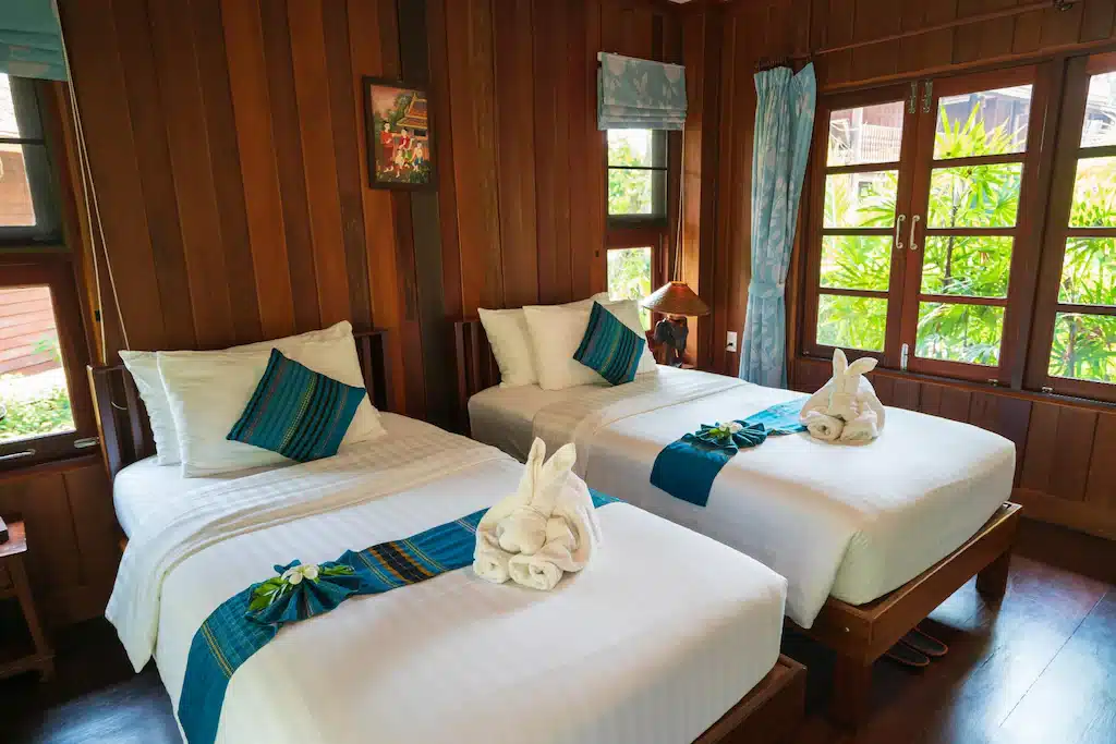 ห้องพักแสนสบายตกแต่งด้วยผนังไม้และเตียง 2 เตียง ตั้งอยู่ในที่พักริมแม่น้ำในจังหวัดราชบุรี ที่พักราชบุรีริมน้ำ