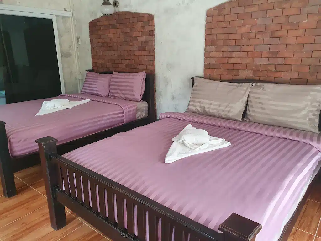 เตียง 2 เตียงพร้อมผ้าปูที่นอนสีม่วงในห้องพักโรงแรมที่มีผนังอิฐ ที่พักริมน้ำราชบุรี