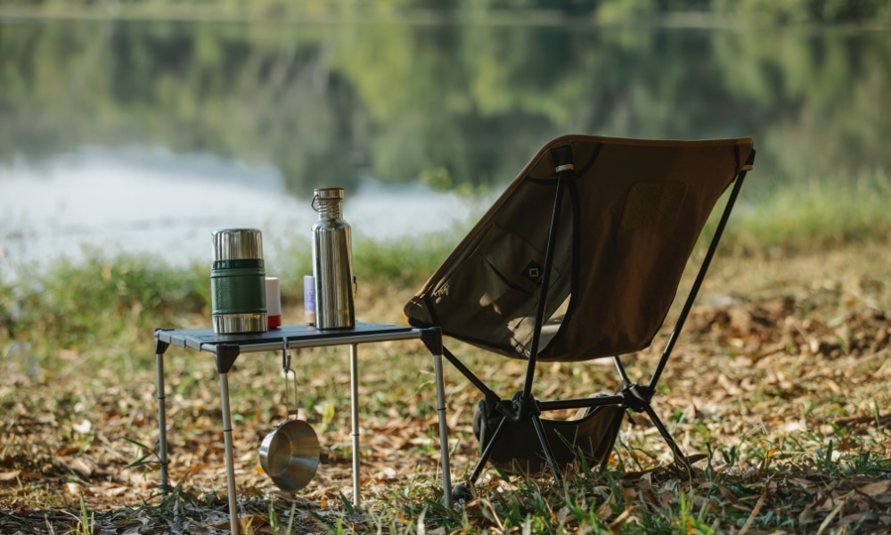เก้าอี้ตั้งแคมป์พร้อมขวดน้ำและถ้วยบนโต๊ะข้างทะเลสาบอันเงียบสงบ กางเต็นท์ดูดาวใกล้กรุงเทพ
