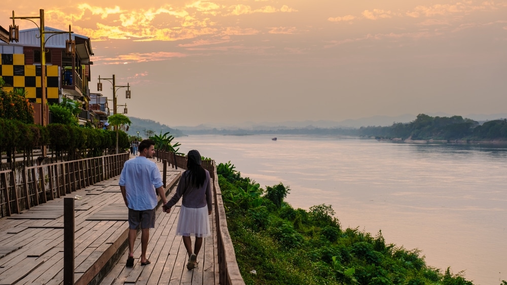 คู่รักเดินไปตามทางเดินริมทะเลใกล้แม่น้ำยามพระอาทิตย์ตกดินในเชียงคาน ที่เที่ยวเดือนตุลาค สถานที่ท่องเที่ยวในจังหวัดเลยของประเทศไทย ที่เที่ยวเชียงคาน
