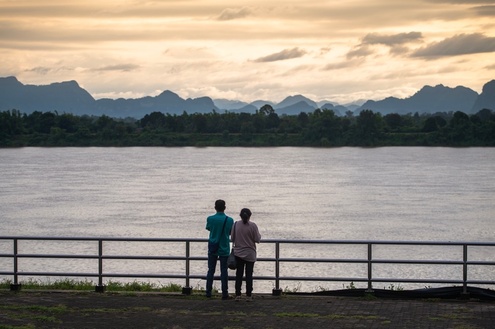 คนสองคนยืนอยู่บนราวบันไดมองออกไปเห็น ที่เที่ยวนครพนม แม่น้ำโดยมีภูเขาเป็นฉากหลังที่เขื่อนศรีนครินทร์