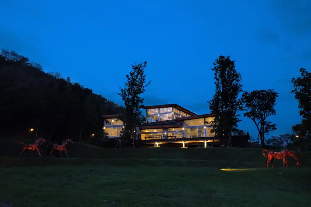 บ้านหลังหนึ่งสว่างไสวในตอนกลางคืนบนเนินเขาหญ้า มองเห็นเมืองชายฝั่งทะเลอันงดงามของชุมพร รีสอร์ทแก่งกระจาน 