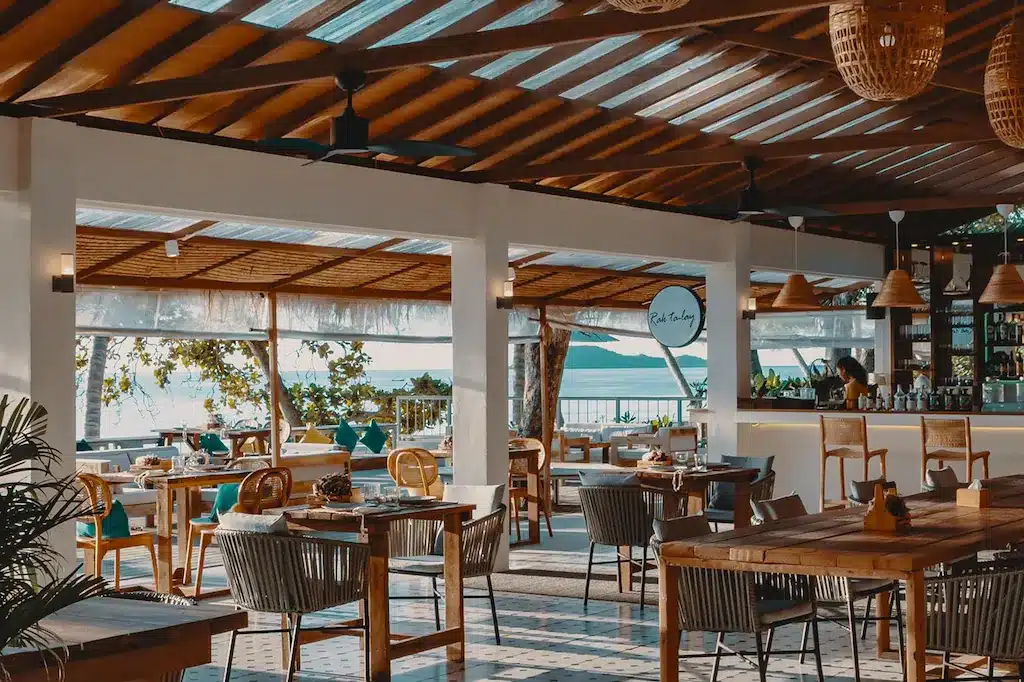 ร้านอาหารที่มีโต๊ะและเก้าอี้ไม้พร้อมวิวทะเล ตั้งอยู่ริมชายหาดที่สวยงามในเมืองชุม ที่พักชุมพรติดทะเล พรอันมีเสน่ห์