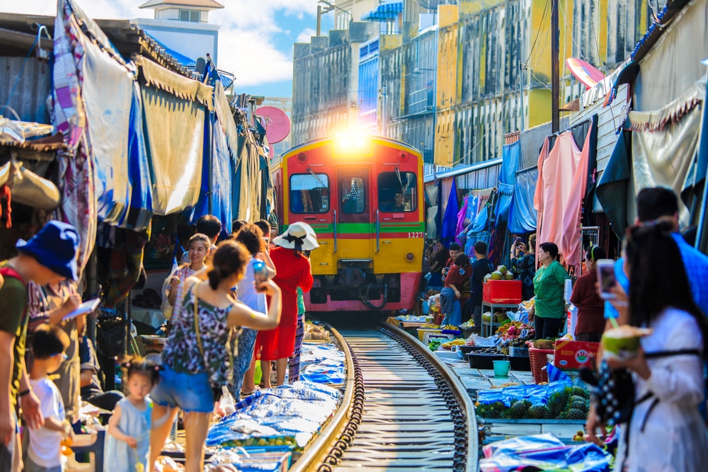 รถไฟแล่นผ่านตลาดที่พลุกพล่าน  ที่เที่ยวสมุทรสงคราม เพียงไม่กี่นิ้วจากแผงลอยและนักช้อปที่เที่ยวสมุทรสงคราม แสงตะวันฉายแสงเจิดจ้า ขณะที่ผู้ชมต่างตั้งตารอการมาถึงอย่างใจจดใจจ่อ.