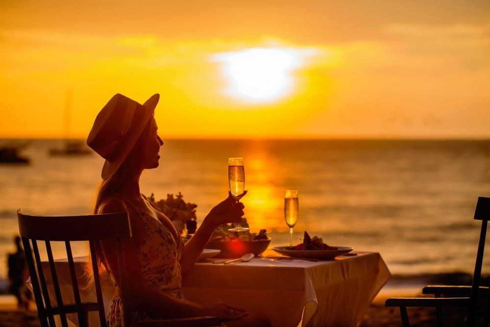 ผู้หญิงคนหนึ่งกำลังนั่งอยู่ที่โต๊ะตอนพระอาทิตย์ตกพร้อมแก้วไวน์ เพลิดเพลินกับบรรยากาศอันเงียบสงบขณะจิบเครื่องดื่มของเธอ