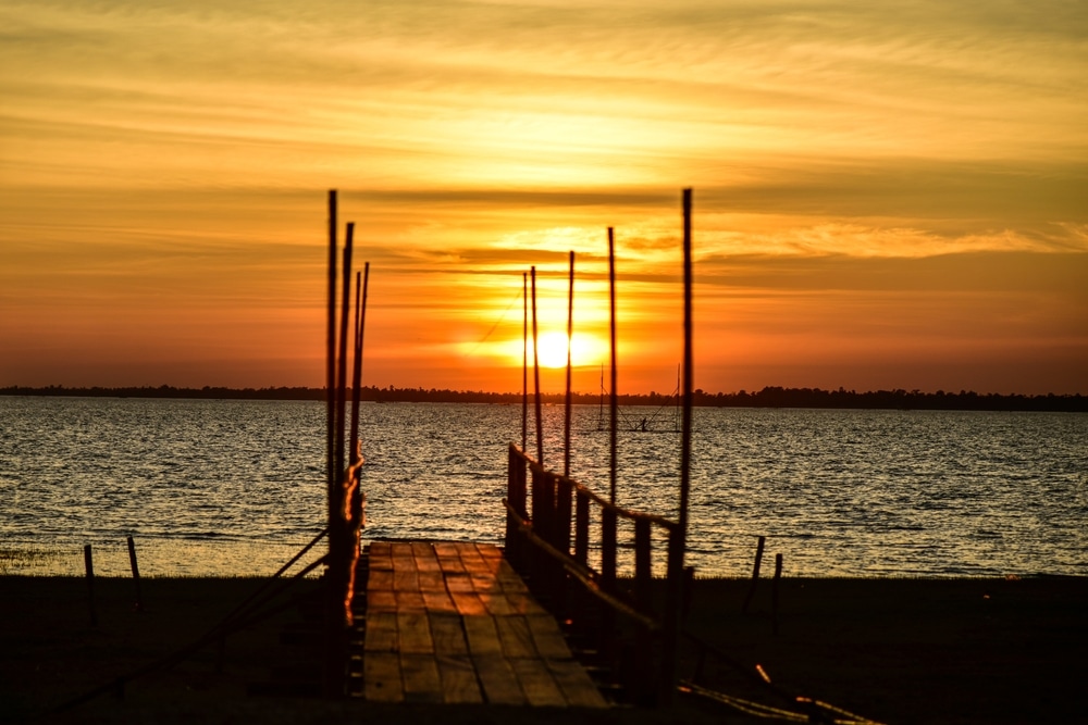 พระอาทิตย์อัสดงเหนือชายหาดมีท่าเทียบเรือไม้และ เขื่อนสิรินธร