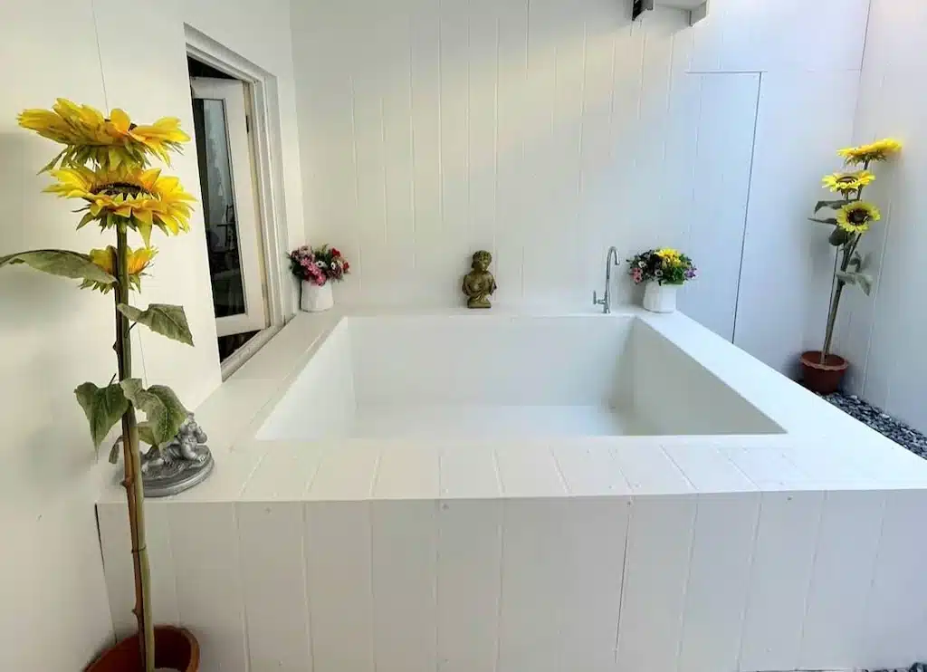 อ่างอาบน้ำสีขาวพร้อมพระพุทธรูปล้อมรอบด้วยดอกไม้ในรีสอร์ทอันเงียบสงบในพื้นที่ท่องเที่ยวน่าน สังขละบุรีที่พัก