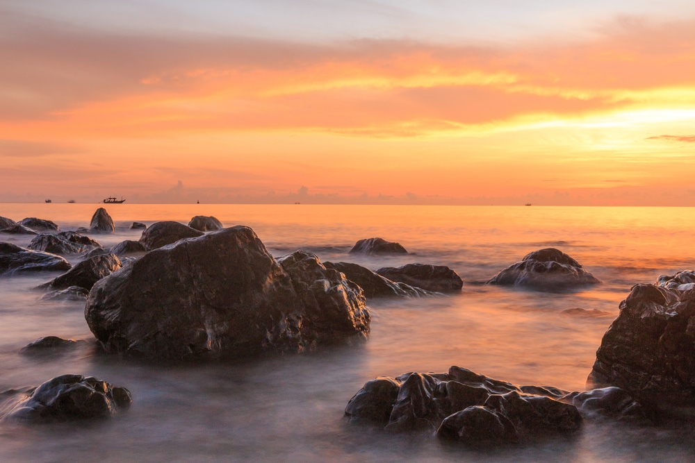 พระอาทิตย์ตกเหนือชายหาดที่มีโขดหินอยู่ในน้ำที่สถานที่ท่องเที่ยวราชบุรี ที่เที่ยวชุมพร