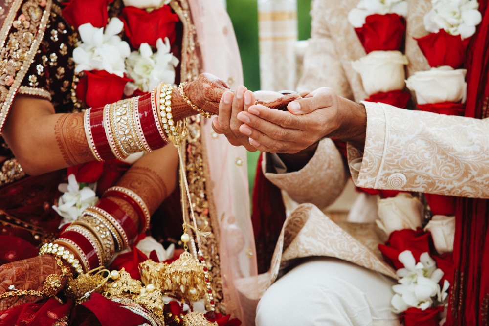 คู่บ่าวสาวแต่งตัวด้วยชุดประจำชาติอินเดียกำลังทำพิธีสวมแหวนในงานแต่งงาน โดยมีดอกไม้ที่มีสีสันสดใสเป็นพื้นหลัง