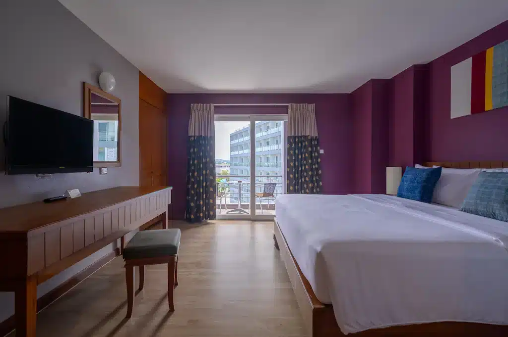 ห้องพักในโรงแรมทันสมัยพร้อมเตียงคู่ ผนังสีม่วง ทางเข้าระเบียงพร้อมทิวทัศน์ทะเลอันตระการตา และโทรทัศน์แบบติดผนัง ที่พักหาดจอมเทียน