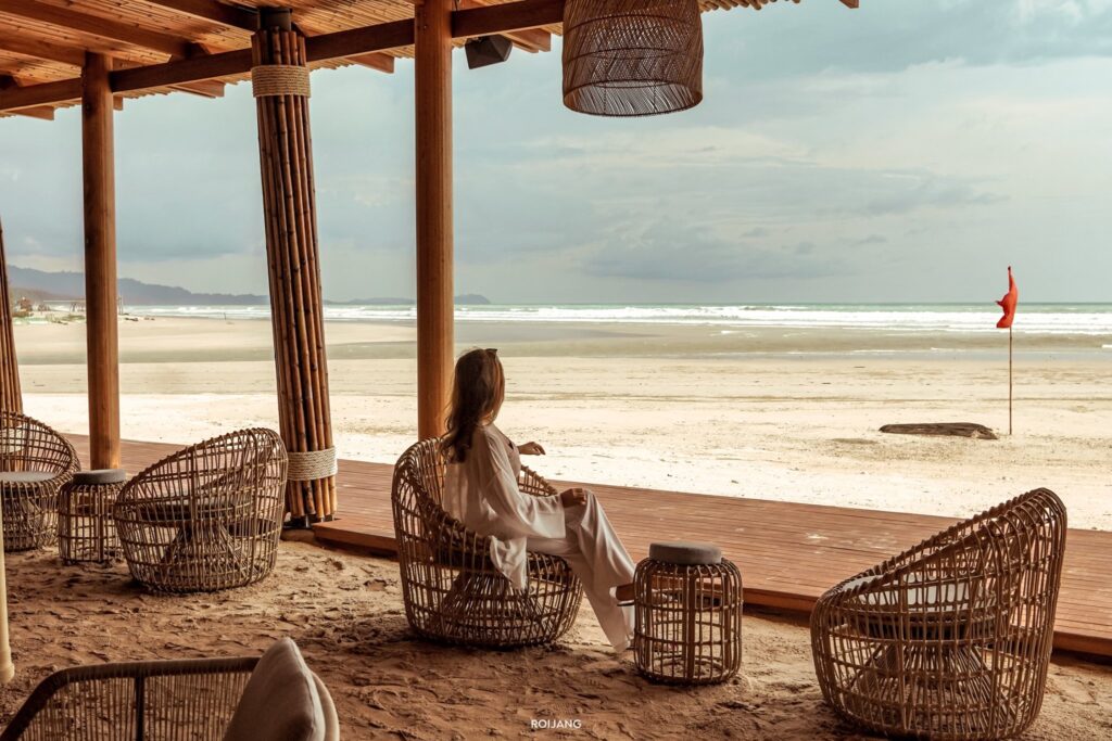 คนที่นั่งบนเก้าอี้หวายใต้ศาลาริมชายหาดในพังงา มองออกไปที่หาดทรายกว้างใหญ่และเส้นขอบฟ้ามหาสมุทร