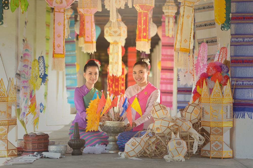 สตรีไทยสองคนในชุดพื้นเมืองกำลังนั่งประดิษฐ์กระถางดอกไม้และประดับธงไตรรงค์เพื่อการตกแต่งงานเทศกาล