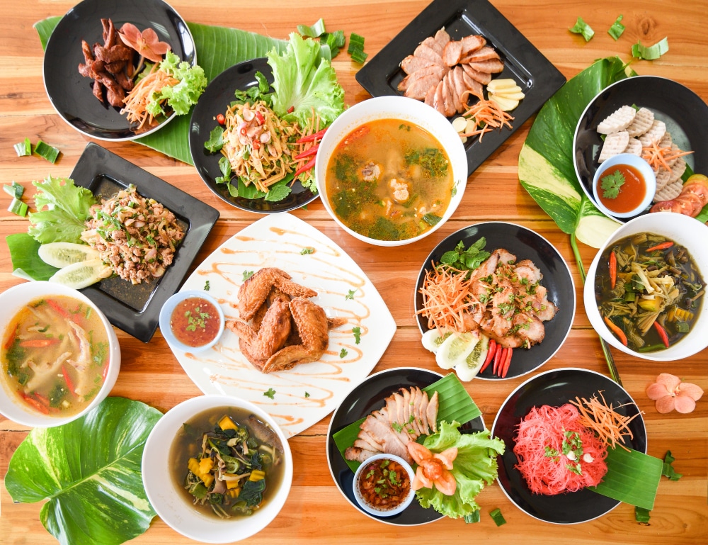 มื้ออาหารไทยที่น่ารับประทานบนโต๊ะไม้ ประกอบด้วยหลากหลายเมนูเช่น ส้มตำ, ลาบ, และต้มยำ