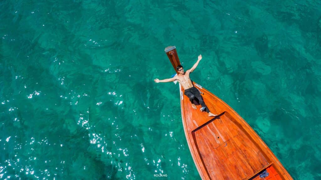 บุคคลยกแขนขึ้นยืนบนหัวเรือไม้ที่ลอยอยู่ในน้ำทะเลสีฟ้าครามใส เที่ยวพังงา