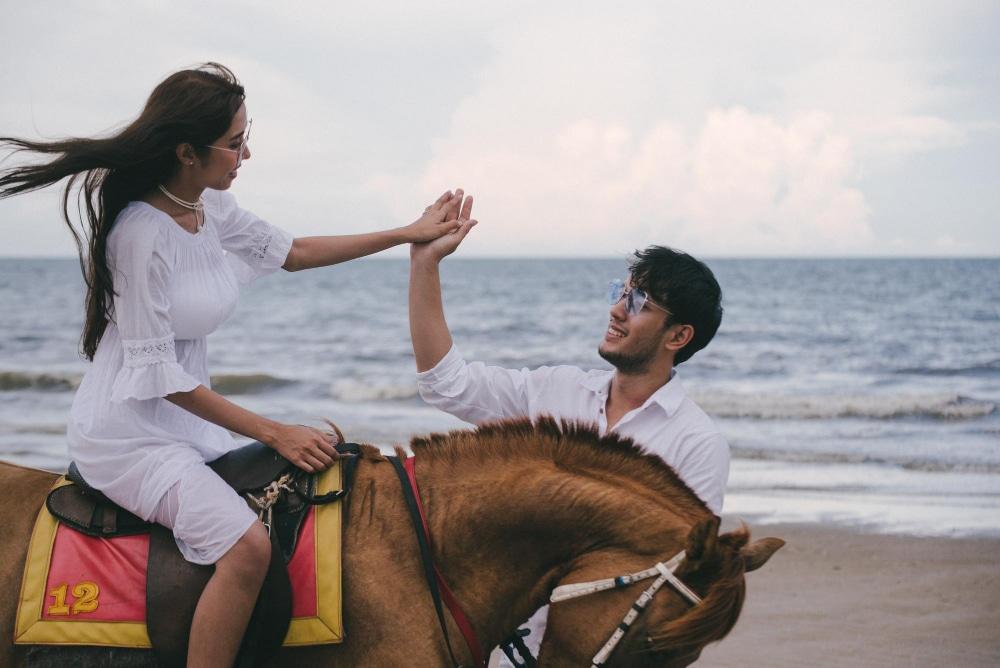 หญิงสาวในชุดขาวกำลังจับมือกับชายหนุ่มที่สวมแว่นกันแดดขณะนั่งบนม้าริมหาดที่สวยงาม
