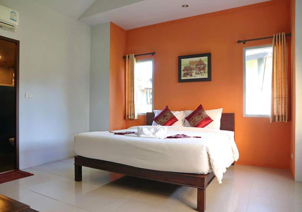 ห้องนอนสว่างสดใสจัดวางอย่างประณีตด้วยผนังสีส้ม เตียงขนาดใหญ่พร้อมผ้าปูเตียงสีขาว  บ้านนาเลาใหม่ หมอนตกแต่ง และทิวทัศน์ของจุดชมวิวดอยหล