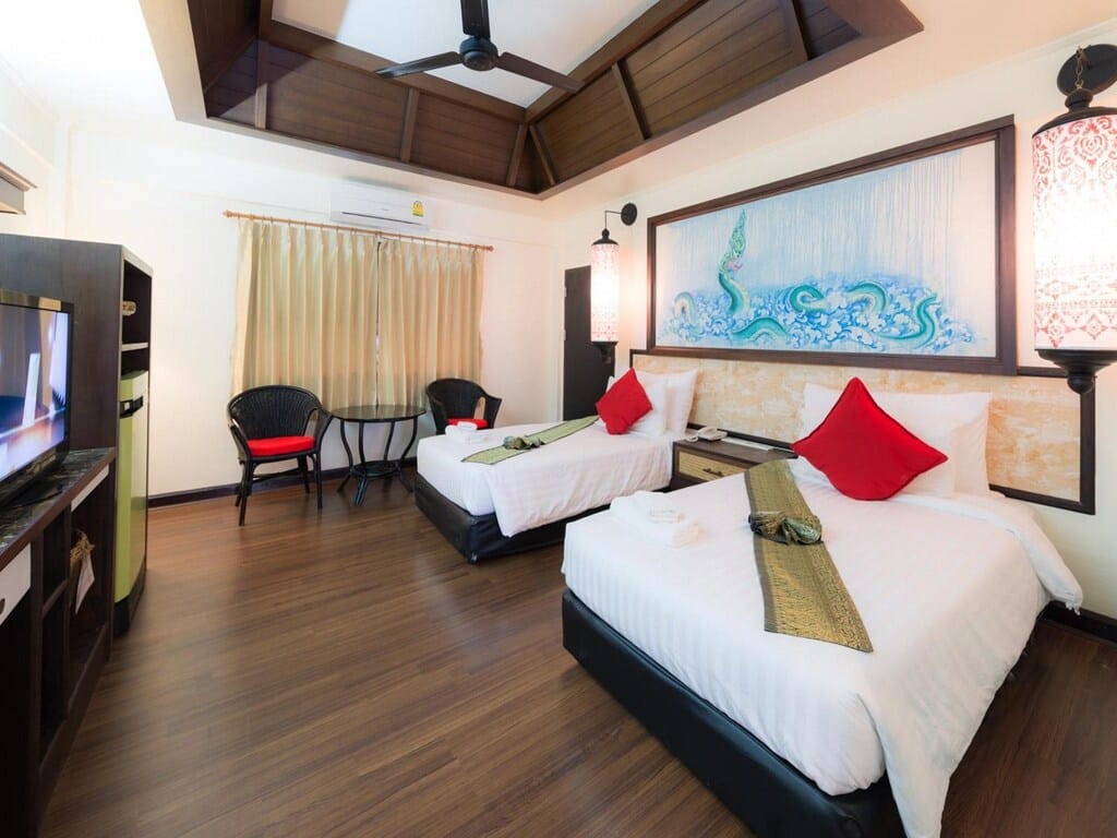 ห้องพักในโรงแรมที่จัดอย่างประณีตในเชียงรายในเมือง มีเตียงคู่ หมอนสีแดง โทรทัศน์ติดผนัง พื้นไม้สีเข้ม และของตกแต่งแบบดั้งเดิม ที่พักในเมืองเชียงราย