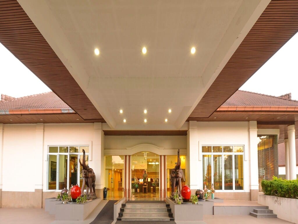 มุมมองที่สมมาตรของทางเข้าโรงแรมที่มีส่วนยื่นกว้าง แสงไฟแบบฝัง และรูปปั้นที่ขนาบข้างประตูกระจก ที่พักมวกเหล็กสระบุรี