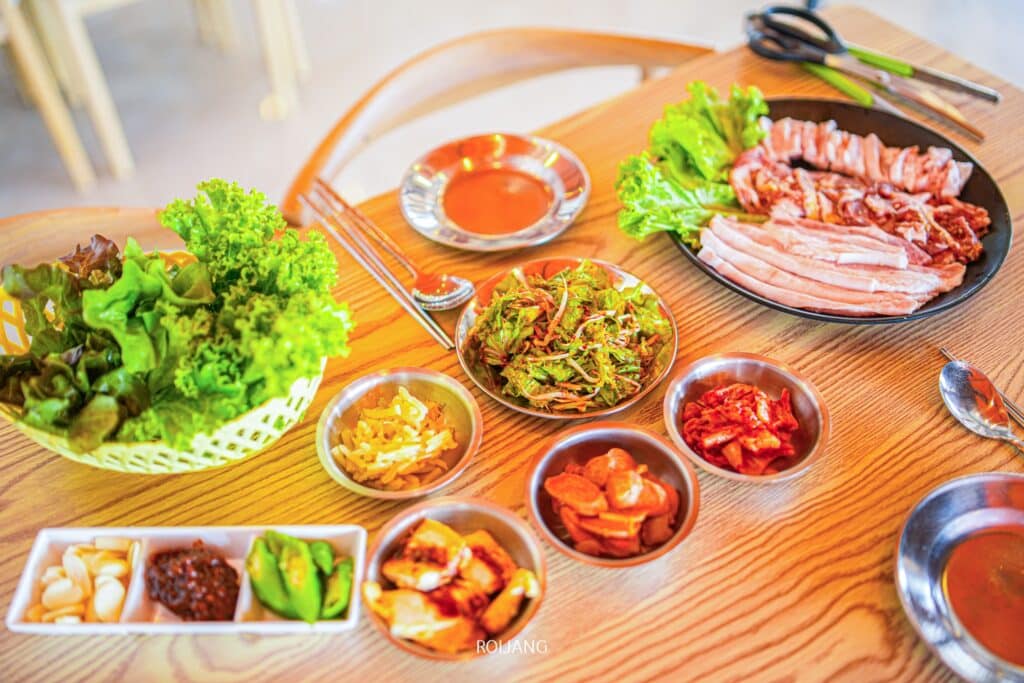 อาหารเกาหลีและเครื่องเคียงหลากหลายชนิดจัดวางอยู่บนโต๊ะไม้ที่ร้านเขาหลัก ประกอบด้วยผักกาดหอมสด หมูสไลซ์ และเมนูต่างๆ ร้านอาหารเขาหลัก