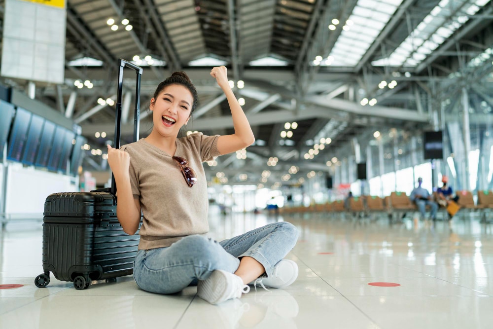 ผู้หญิงคนหนึ่งนั่งยิ้มแย้มอยู่ข้างกระเป๋าเดินทางที่สนามบิน แสดงท่าทีที่มีความสุขและตื่นเต้นกับการเดินทางของเธอ "ทำไมคนอินเดียเลือกแต่งงานที่ประเทศไทย?"