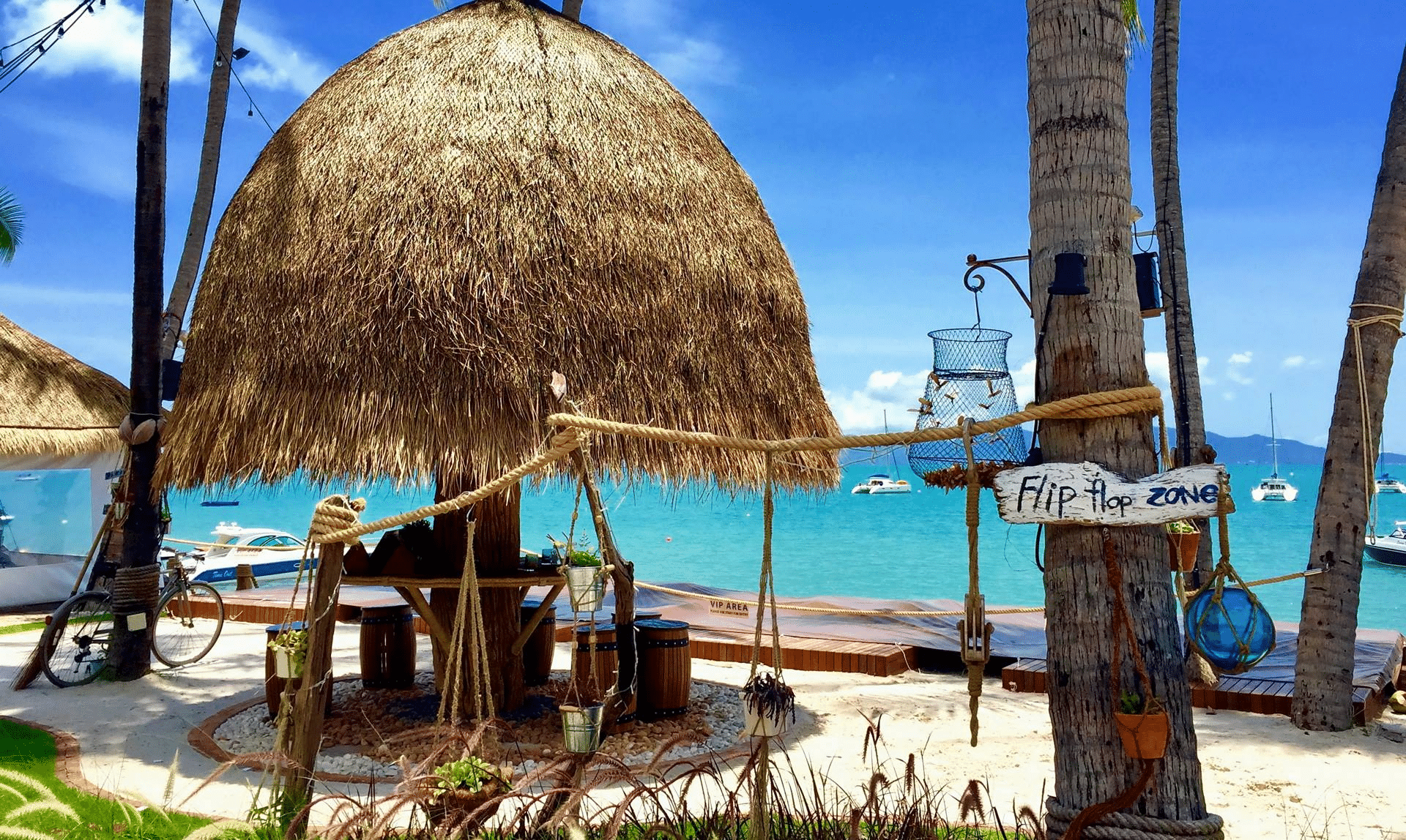 กระท่อมชายหาดมุงจากที่มีรั้วไม้และป้าย "Flip Flop Zone" ต้นปาล์มและ ที่เที่ยวเกาะสมุย เรือที่ทอดสมออยู่บนท้องทะเลสีฟ้าใสภายใต้ท้องฟ้าสีฟ้า เหมาะสำหรับผู้ที่ต้องการเที่ยวเกาะสมุยและดื่มด่ำกับบรรยากาศสบายๆ ของเกาะ.