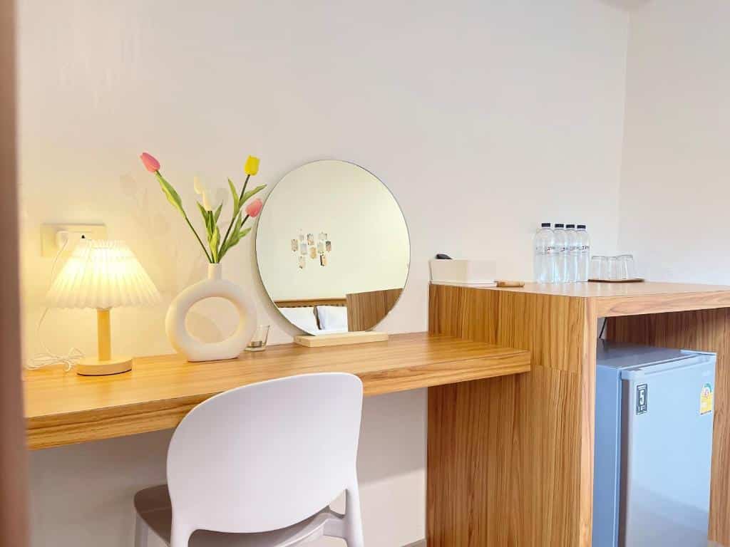มุมห้องที่มีแสงสว่างเพียงพอพร้อมโต๊ะเครื่องแป้งไม้ กระจกทรงกลม เก้าอี้สีขาว โคมไฟ แจกันดอกไม้ และตู้เย็นขนาดเล็ก ที่พักเบตง