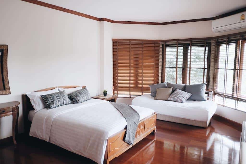 ห้องนอนแสนสบายที่มีแสงสว่างเพียงพอพร้อมเตียง 2 เตียงที่จัดอย่างประณีตพร้อมโซฟาใกล้หน้าต่าง พื้นไม้ และมู่ลี่ ที่พักมวกเหล็ก