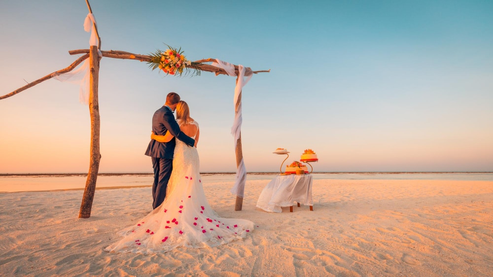 งานแต่งงานบนชายหาดที่โรแมนติกตอนพระอาทิตย์ตกดิน มีโต๊ะแต่งงานตกแต่งด้วยดอกไม้สดและลูกโป่งที่สวยงามตั้งอยู่บนทราย